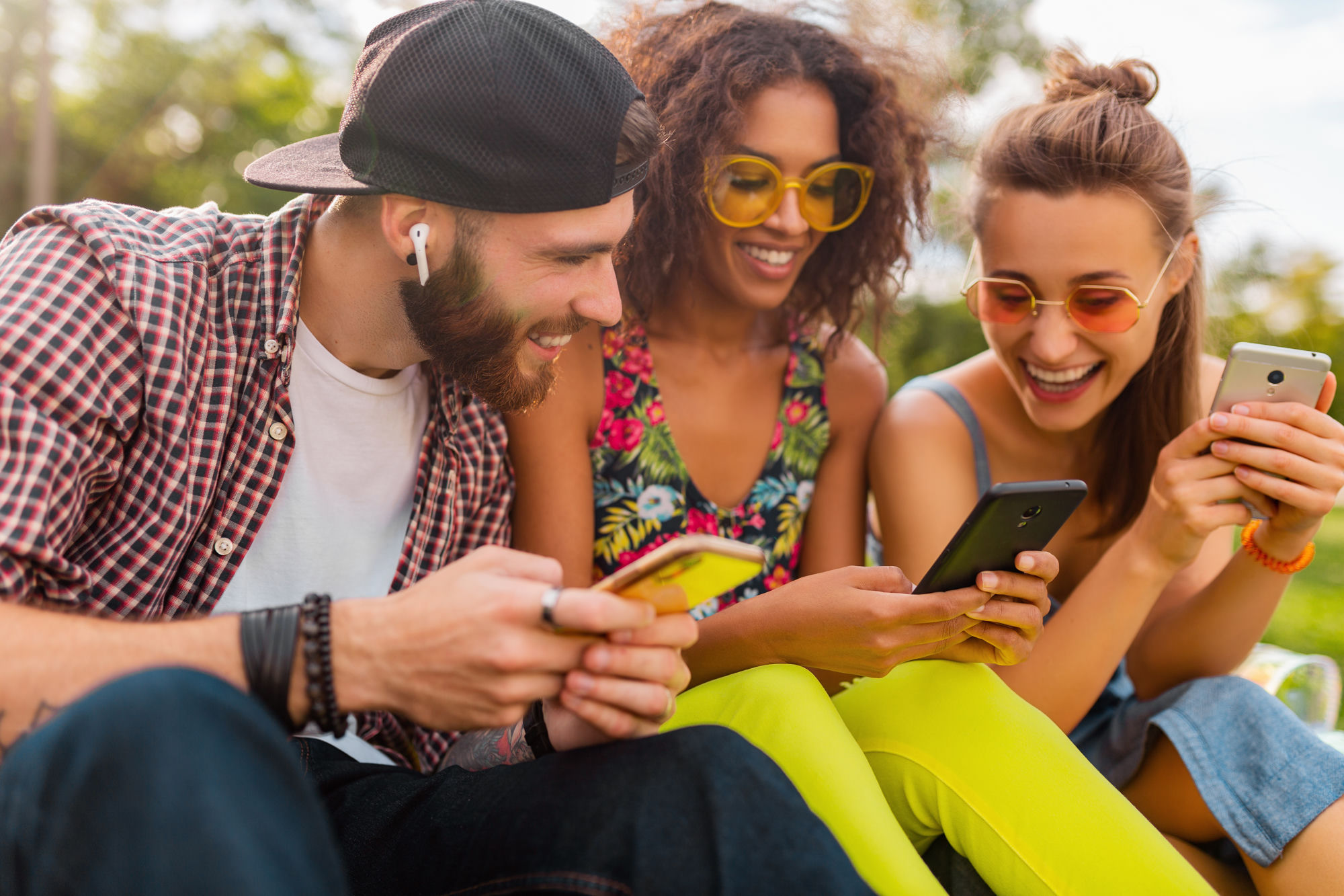 Three friends laughing and looking at their phones together outdoors