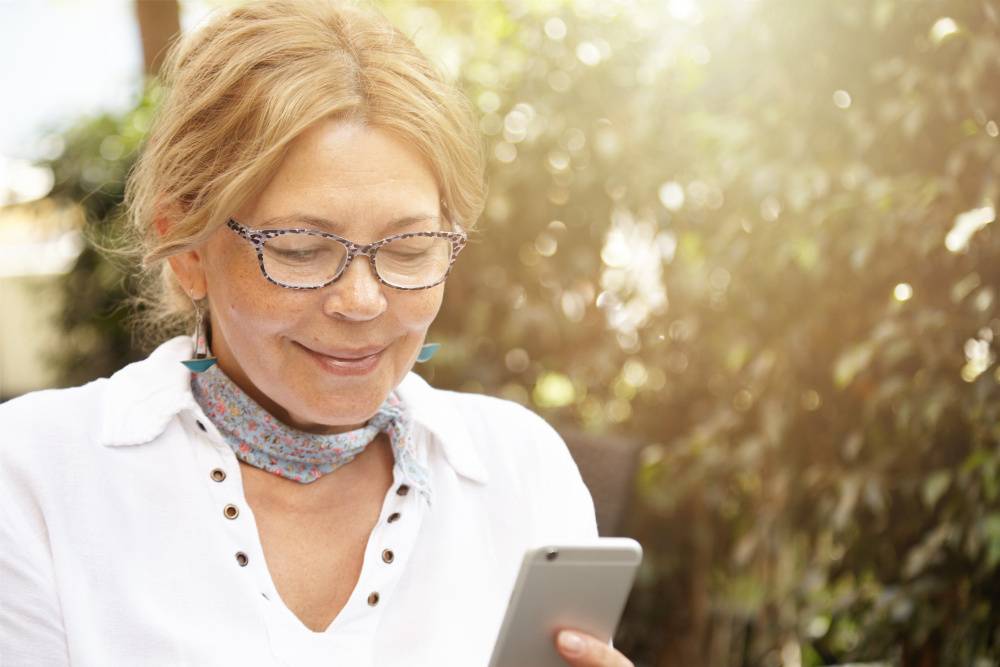 Smiling older woman with glasses looking at her phone outdoors