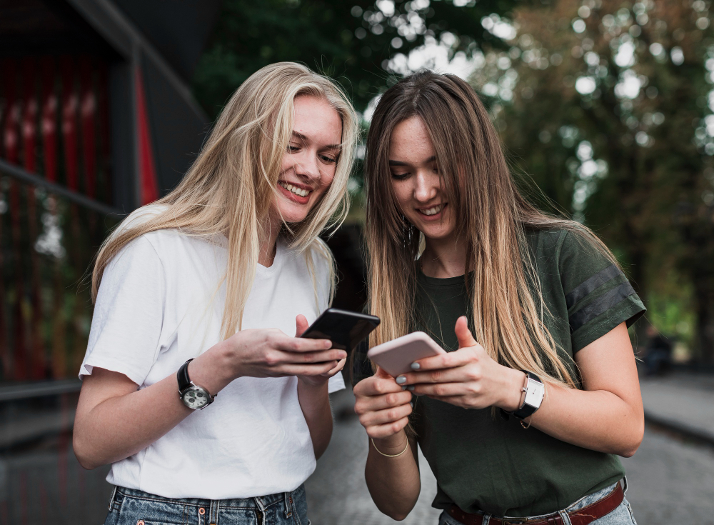 Two smiling young women looking at their phones together outdoors