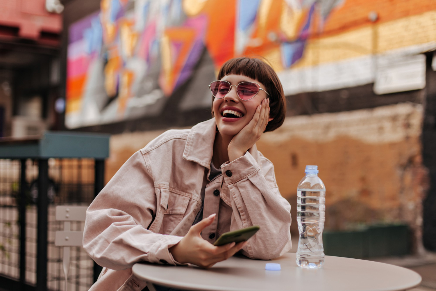 Laughing woman in sunglasses sitting at an outdoor cafe table with her phone