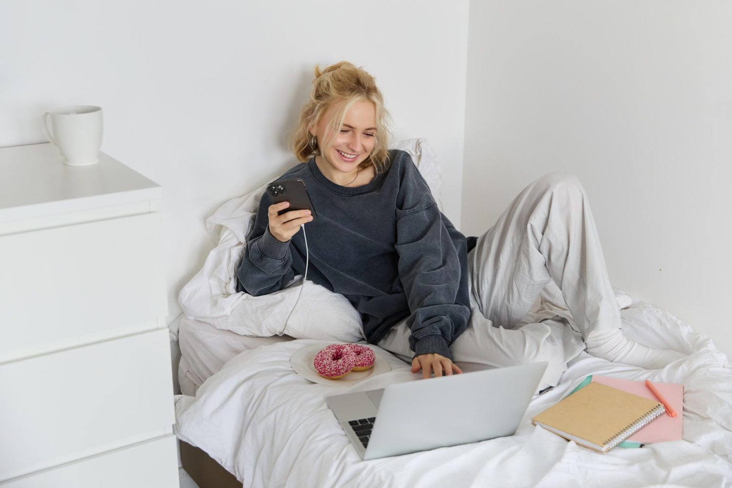 Smiling woman sitting on her bed with a laptop, phone, and donuts