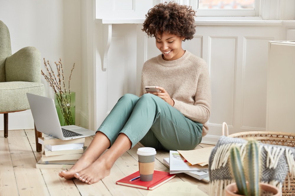 Smiling woman with curly hair sitting on the floor looking at her phone next to a laptop and coffee