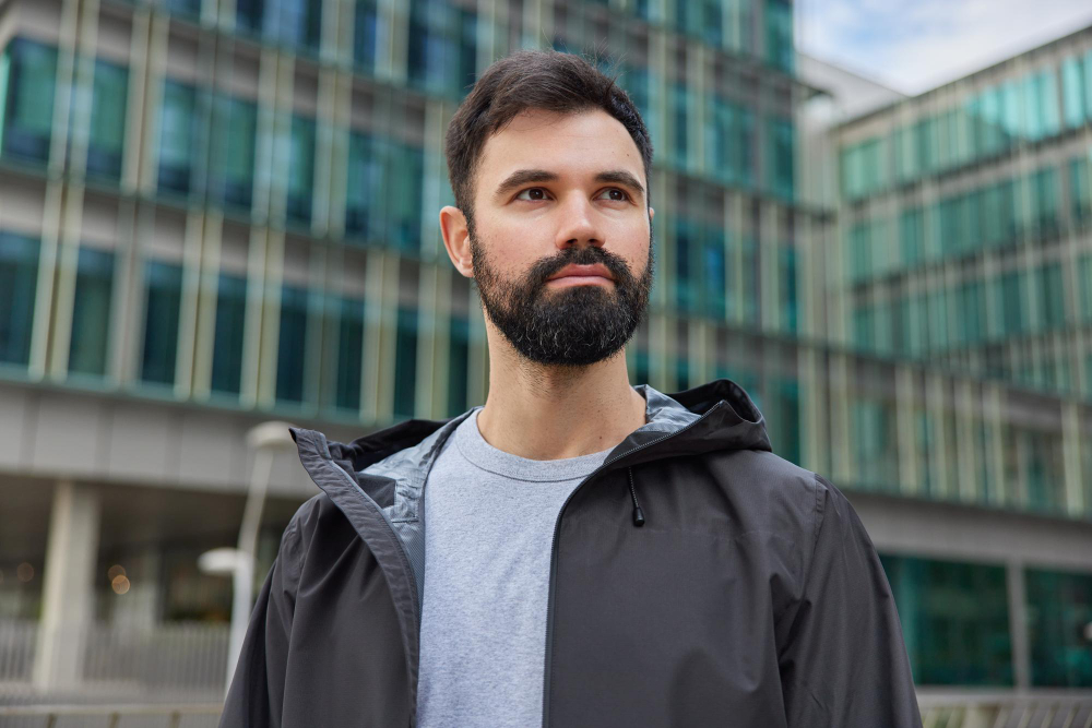 Man with a beard looking off to the side on a city street lined with glass buildings
