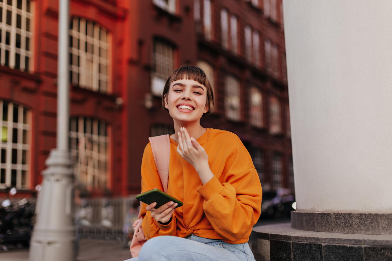 Smiling woman in an orange sweater holding her phone outdoors in the city