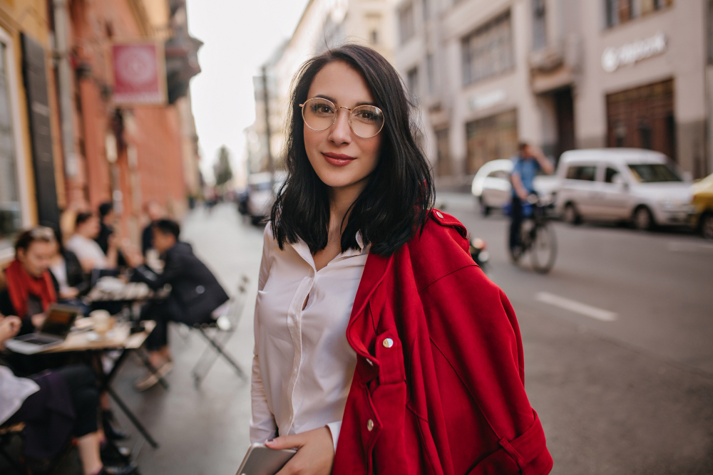 Smiling woman in glasses and a red jacket standing on a city street