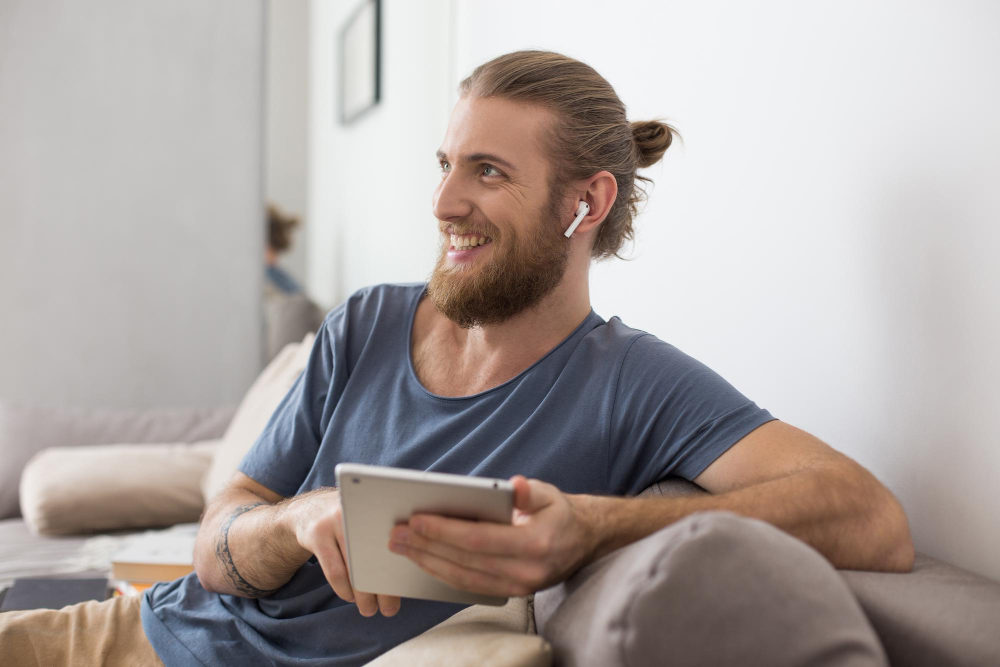 Smiling man with wireless earbuds relaxing on a couch holding a tablet